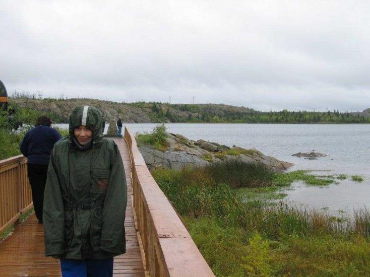 Personne marchant sur la promenade de bois mouillé de Flin Flon avec son imperméable et son capuchon relevé après une averse de Dame Nature.