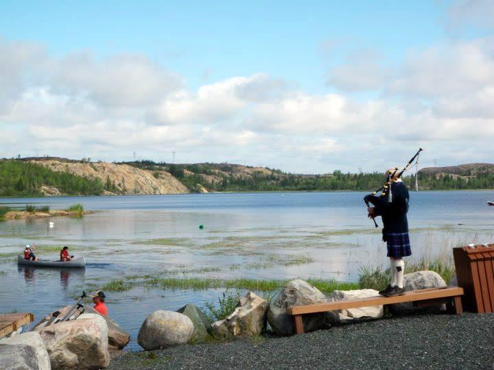 Un joueur de cornemuse sur un banc surplombe les eaux tandis que des canoéistes glissent le long des rives.