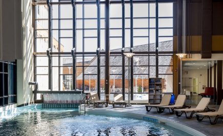 Des chaises longues sont alignées le long d'une piscine, devant un mur de baies vitrées donnant sur l'extérieur.