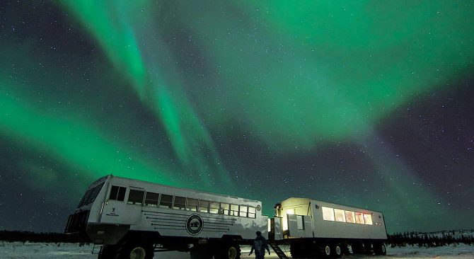 Une personne se tenant dans la toundra devant les véhicules du Dan's Diner sous une aurore boréale.