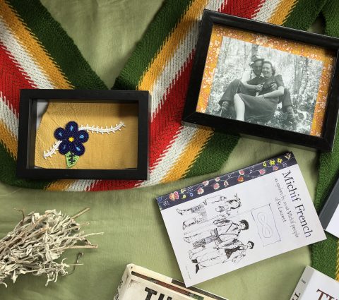 Une table sur laquelle sont posés divers objets - une photo encadrée en noir et blanc, une fleur en perles encadrée, une écharpe métisse et un livre.