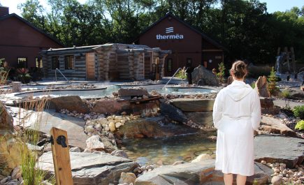 Une dame en robe blanche surplombant les piscines extérieures et le spa de Thermëa by Nordik Spa-Nature.